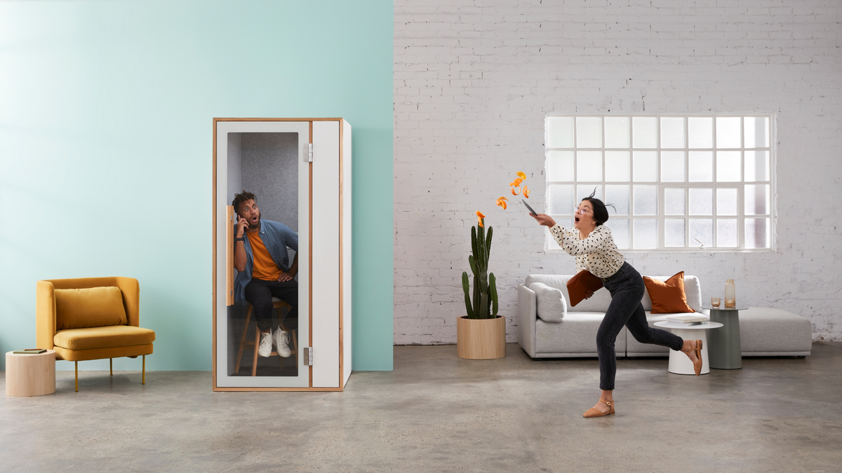 Man calmly on a phone call inside a soundproof office phone booth, while a woman juggles fire in a chaotic space beside it